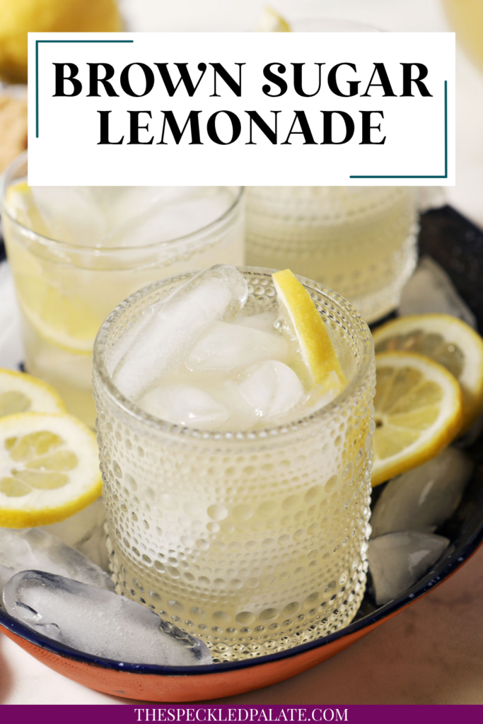 Four glasses of brown sugar lemonade on a blue tray with ice cubes and lemon wheels on a marble surface, next to a blue patterned linen, more lemon slices, and a bowl of brown sugar with the text "Brown Sugar Lemonade" on top.