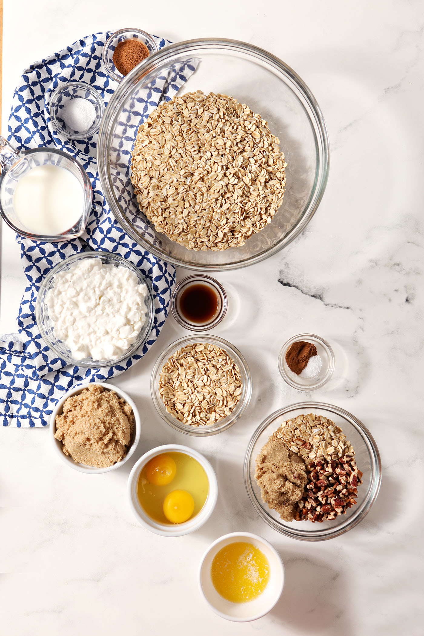 Ingredients to make baked oatmeal cups in bowls on a marble surface with a patterned blue linen