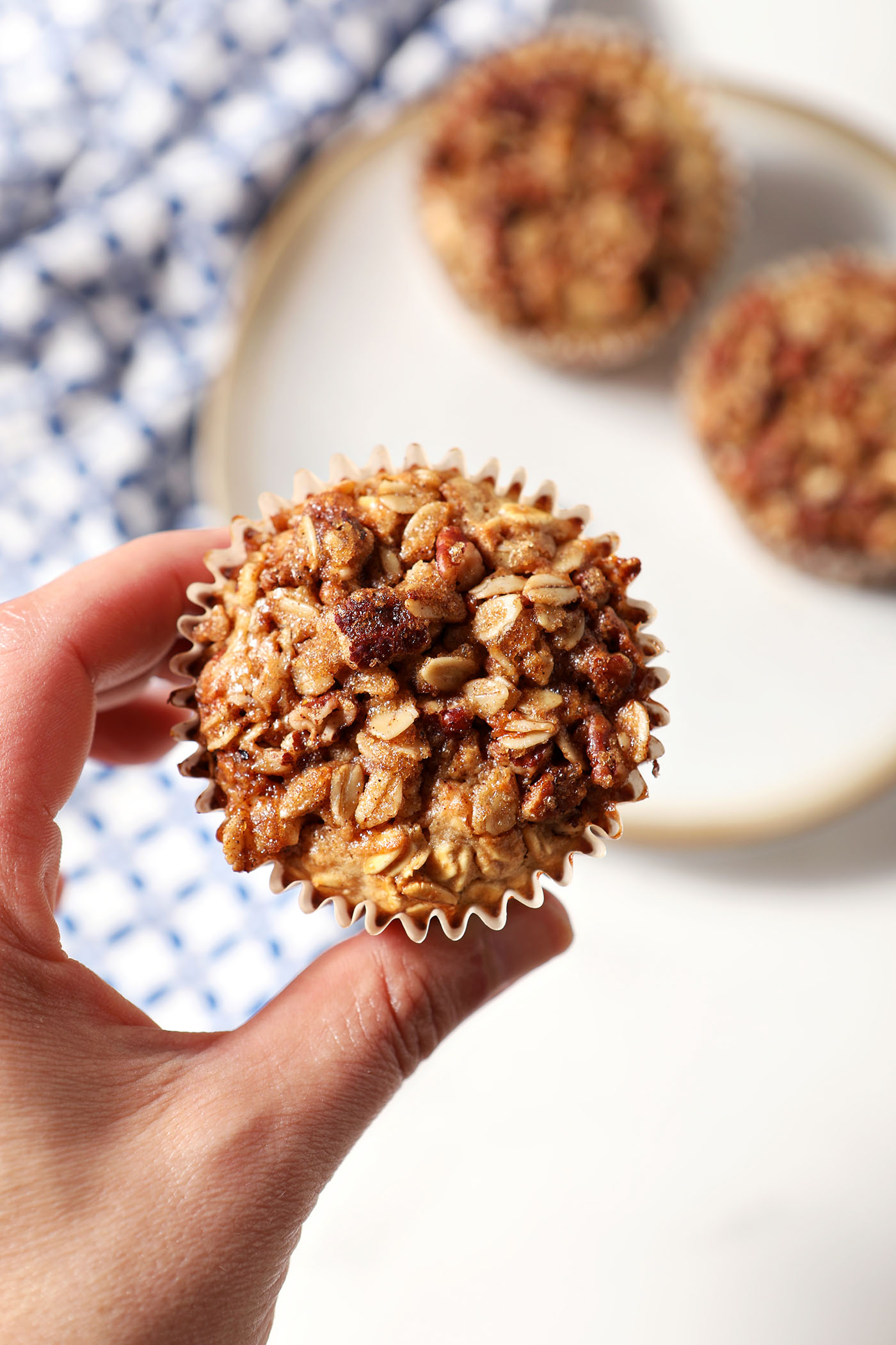 A hand holds an oatmeal muffin cup in hand above a plate with two more and a blue patterned linen