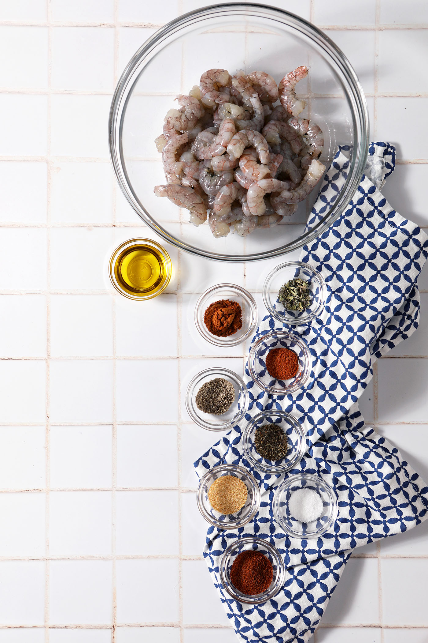 Ingredients to make blackening seasoning for shrimp in bowls with shrimp tails and oil on a white tiled surface with a blue linen