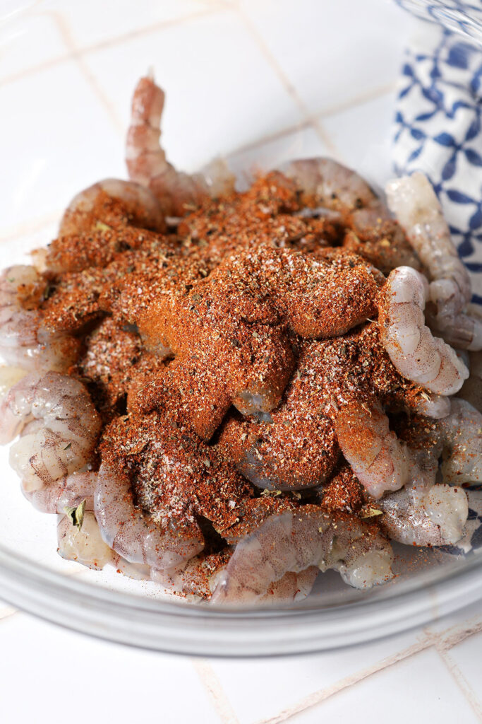 Blackening seasoning on top of raw shrimp in a bowl on a white tiled surface next to a patterned blue linen