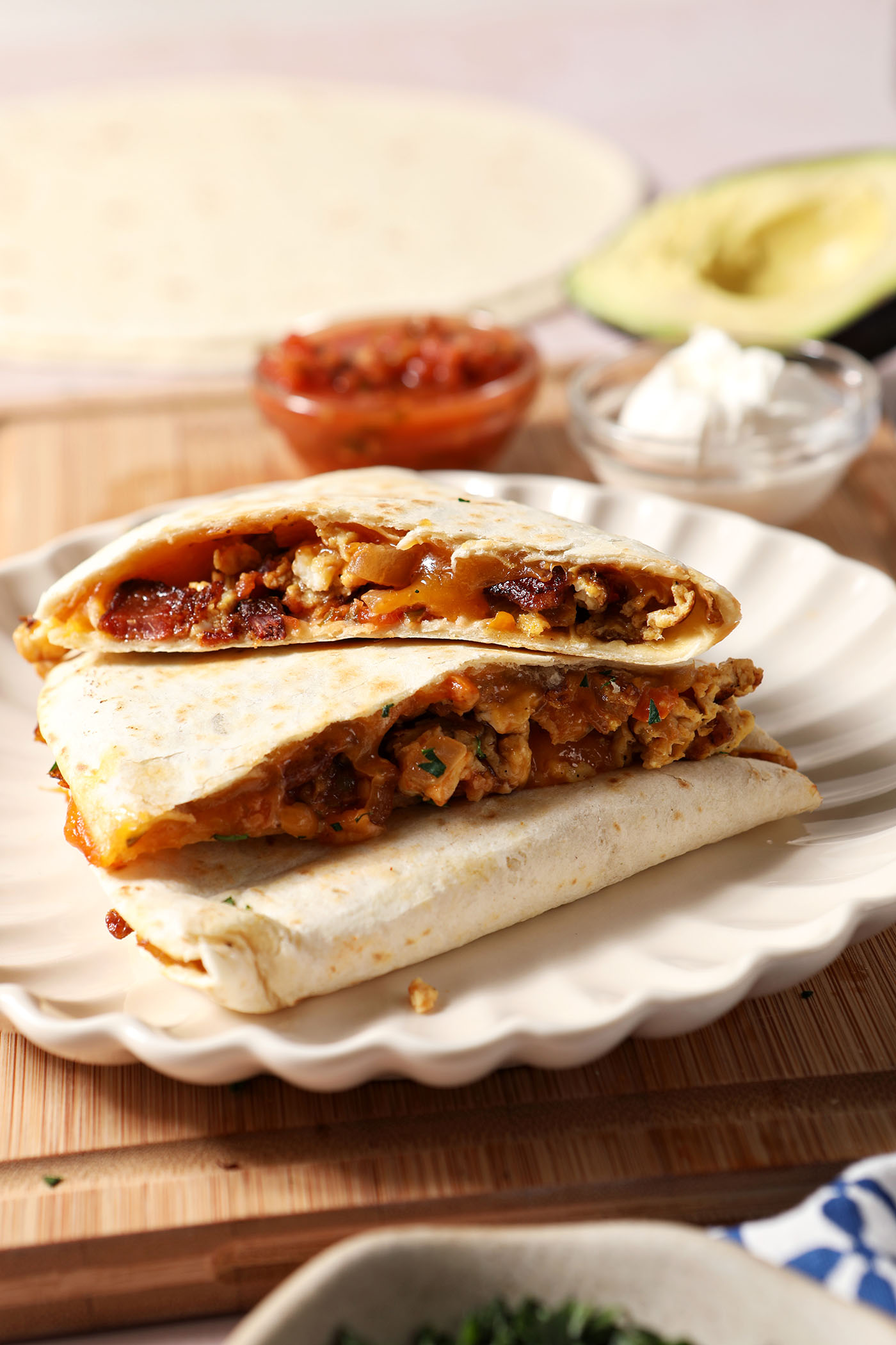 Breakfast quesadillas stacked on a plate on top of a wooden cutting board next to bowls of salsa and sour cream and an avocado.