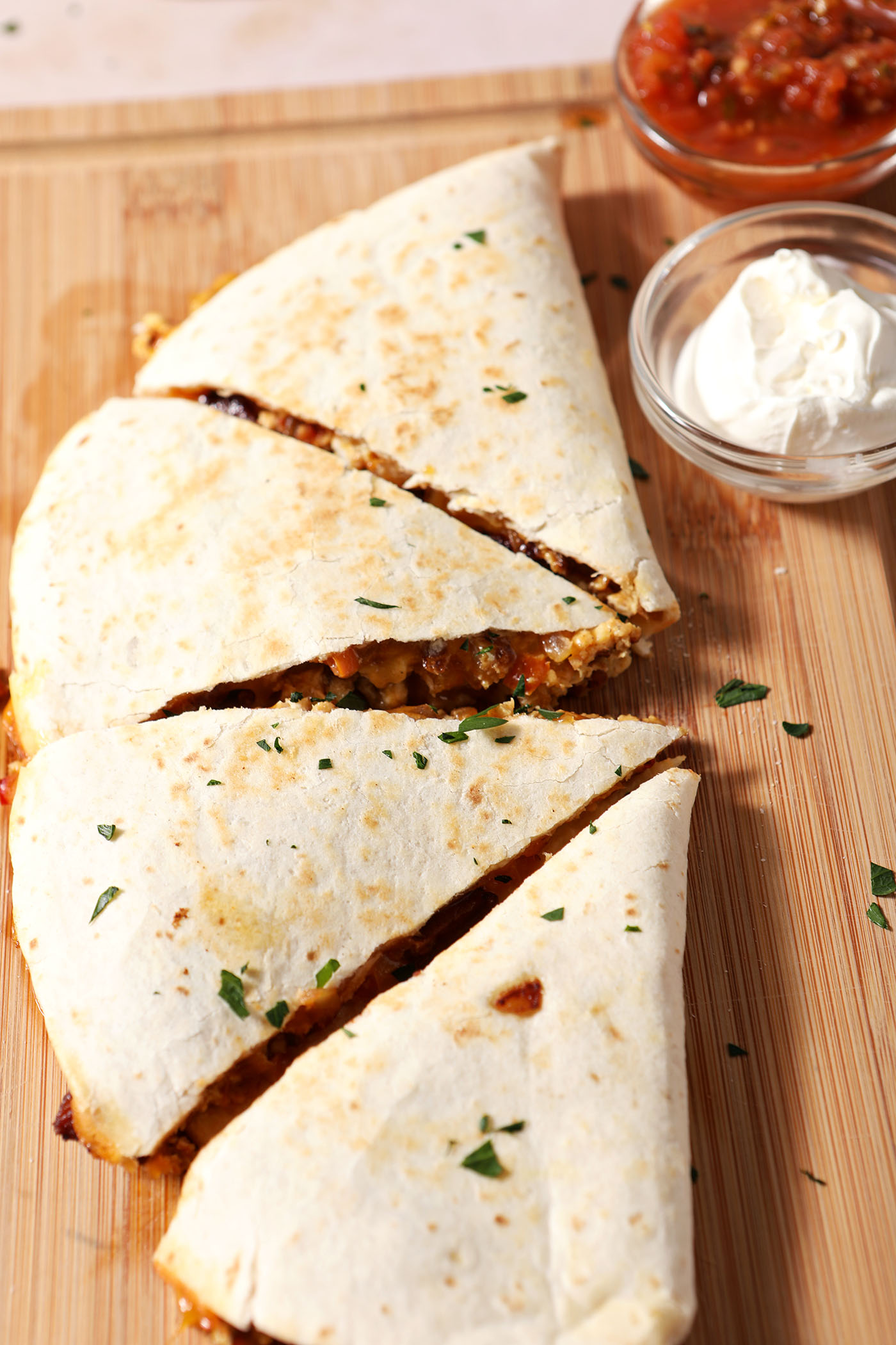 A sliced quesadilla on a wooden cutting board next to bowls of salsa and sour cream.