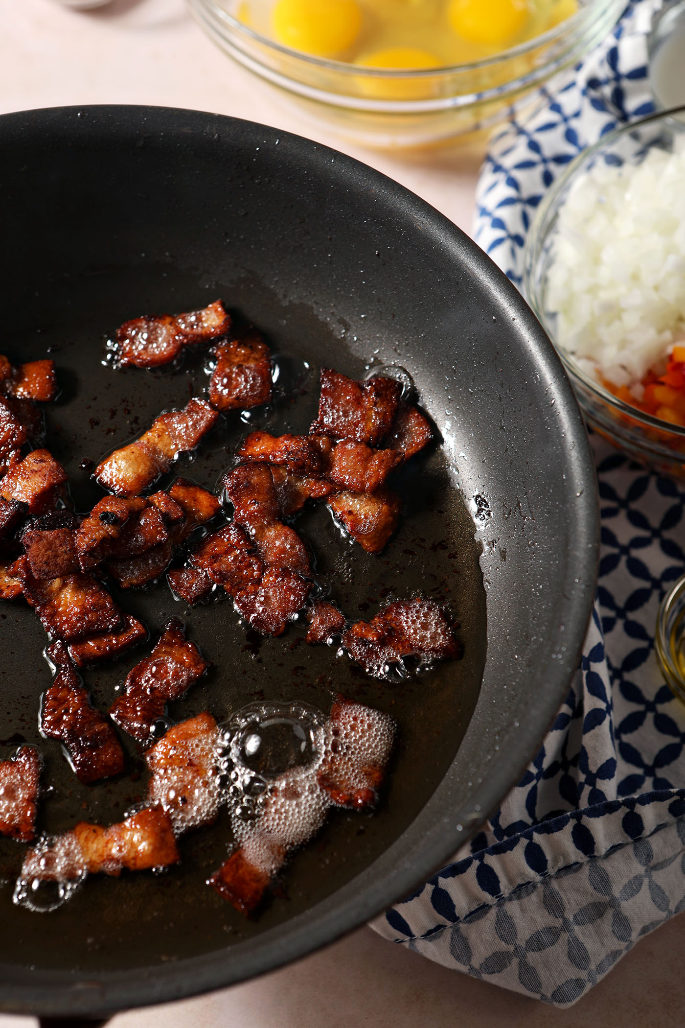 Bacon fries in a skillet next to bowls of other ingredients