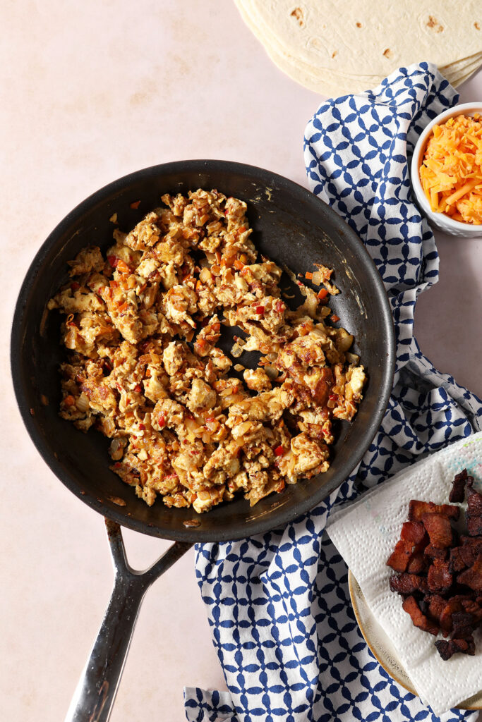 Scrambled eggs in a skillet, surrounded by cooked bacon, shredded cheese and tortillas, with a blue patterned linen