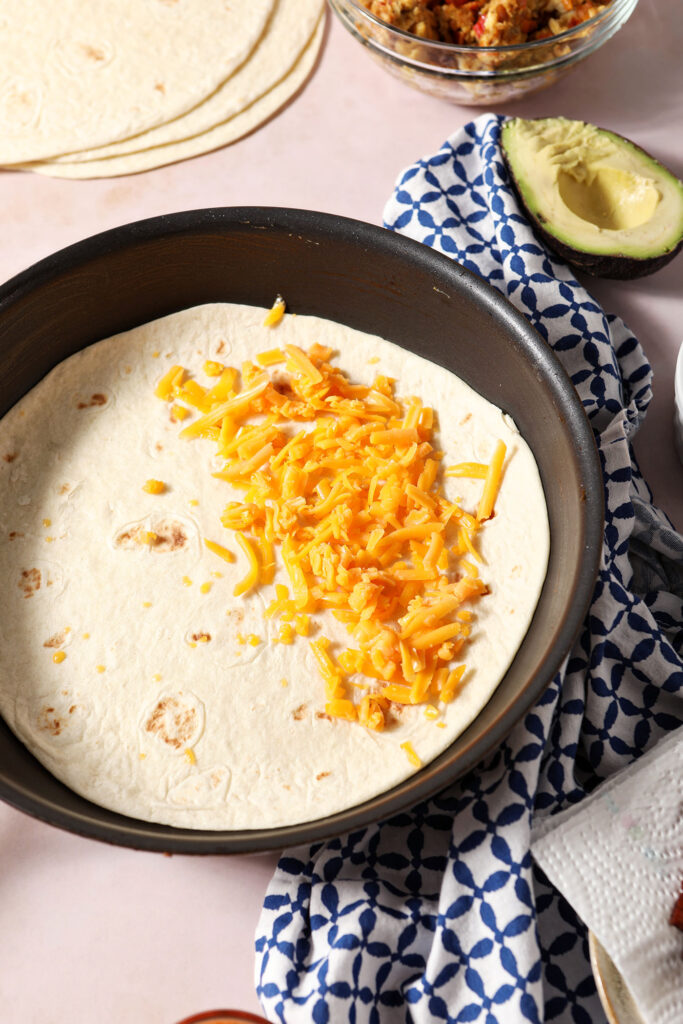A tortilla in a skillet with shredded cheddar cheese over half of it