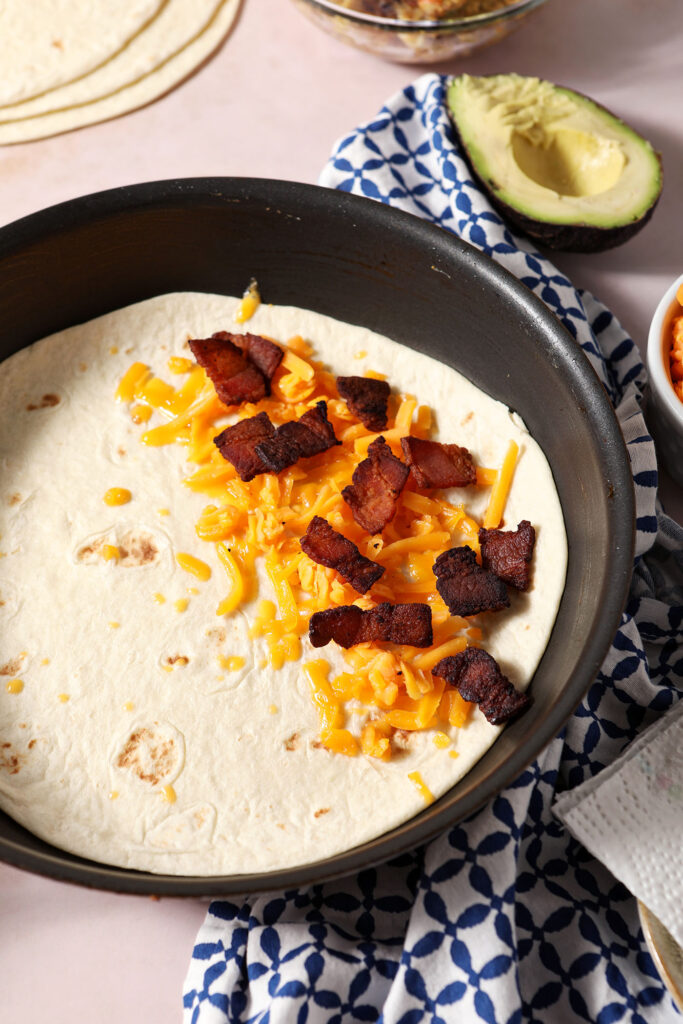 Chopped cooked bacon on top of shredded cheddar cheese on top of a tortilla in a skillet