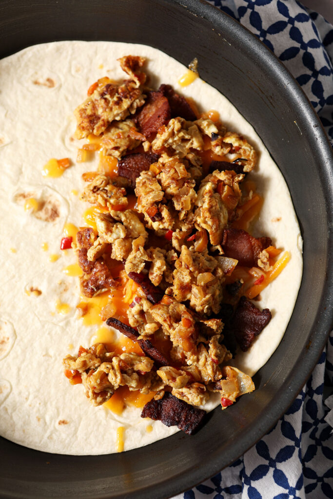 Scrambled eggs on top of the bacon and cheese mixture on a tortilla in a skillet