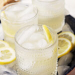 Three lemonades sit on a blue tray surrounded by ice cubes and sliced lemons