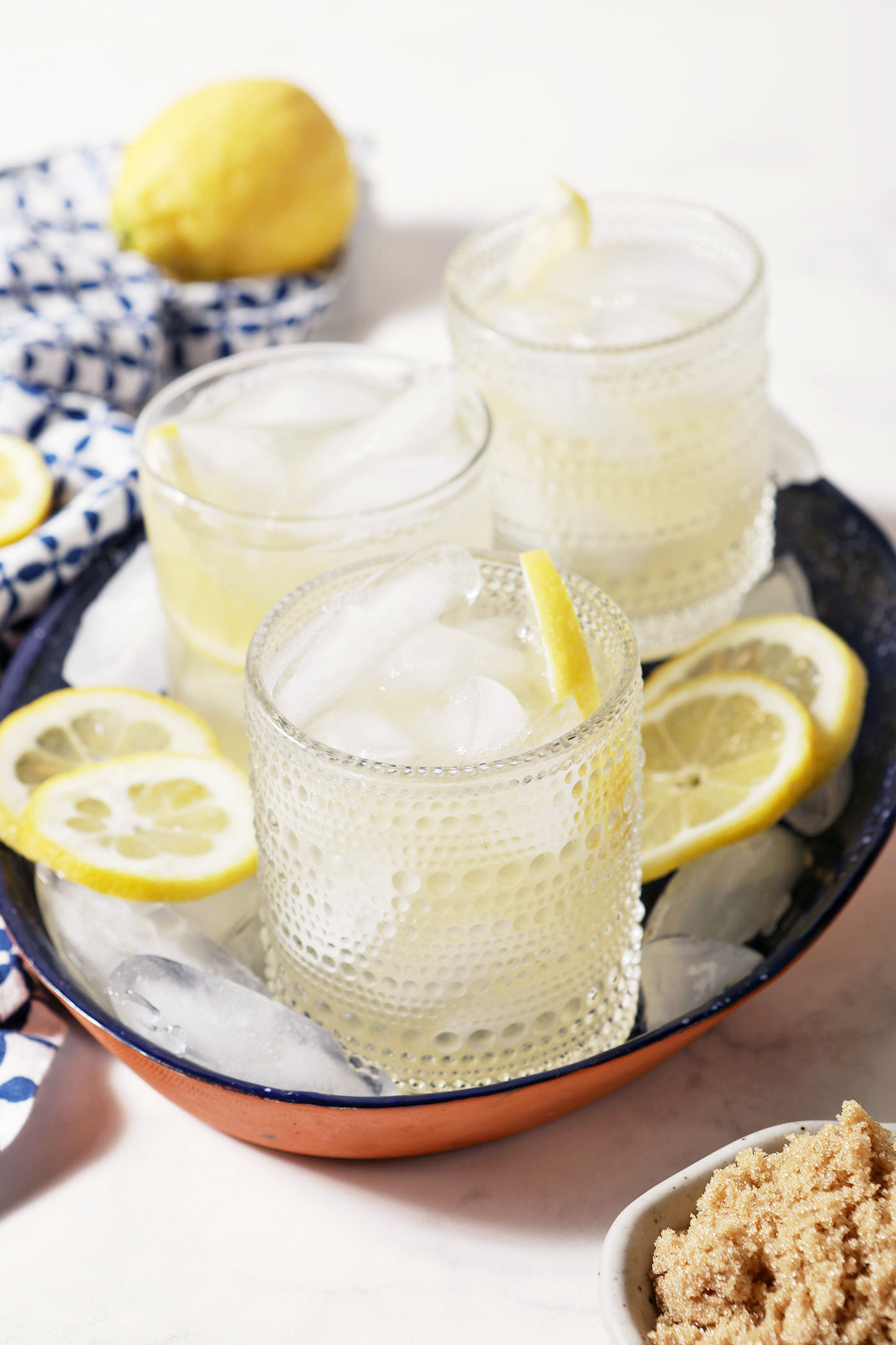 Three glasses of brown sugar lemonade on a blue tray, surrounded by ice cubes, lemon slices, and a blue patterned linen