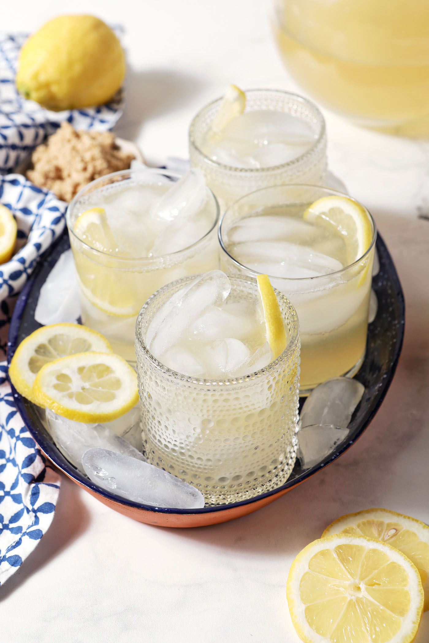 Four glasses of brown sugar lemonade on a blue tray with ice cubes and lemon wheels on a marble surface, next to a blue patterned linen, more lemon slices, and a bowl of brown sugar