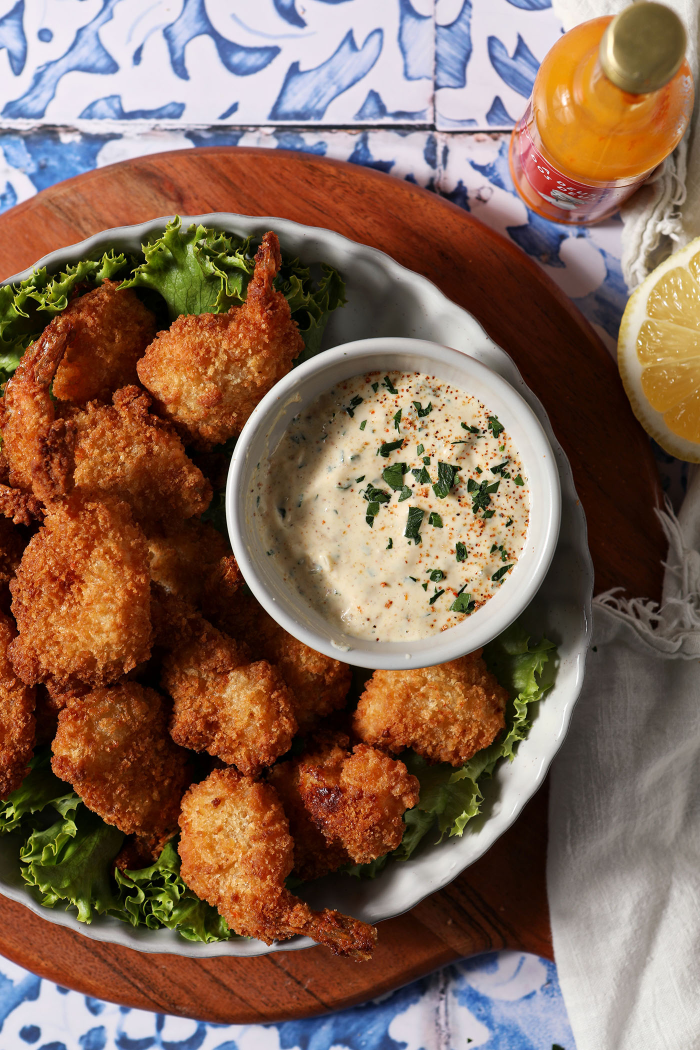A bowl of Cajun Remoulade sauce sits on a platter with fried shrimp and fresh lettuce on top of a wooden platter on a blue and white tile surface.
