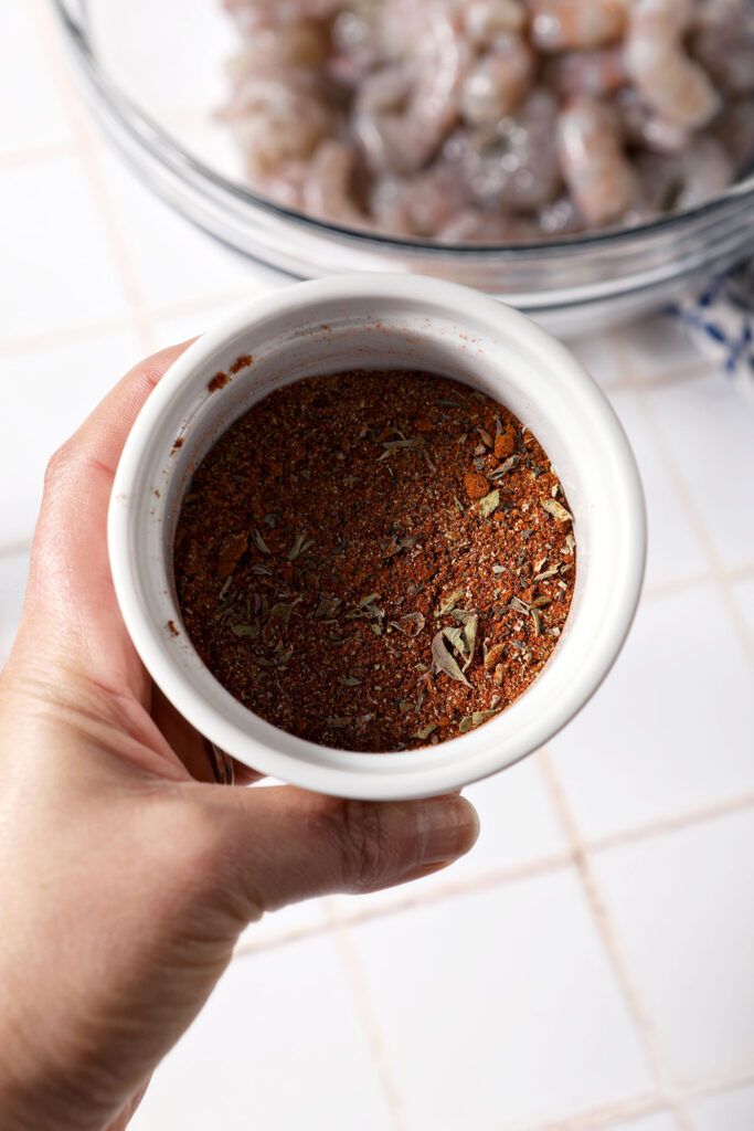 Blackening seasoning in a white container held in a hand above a bowl of raw shrimp