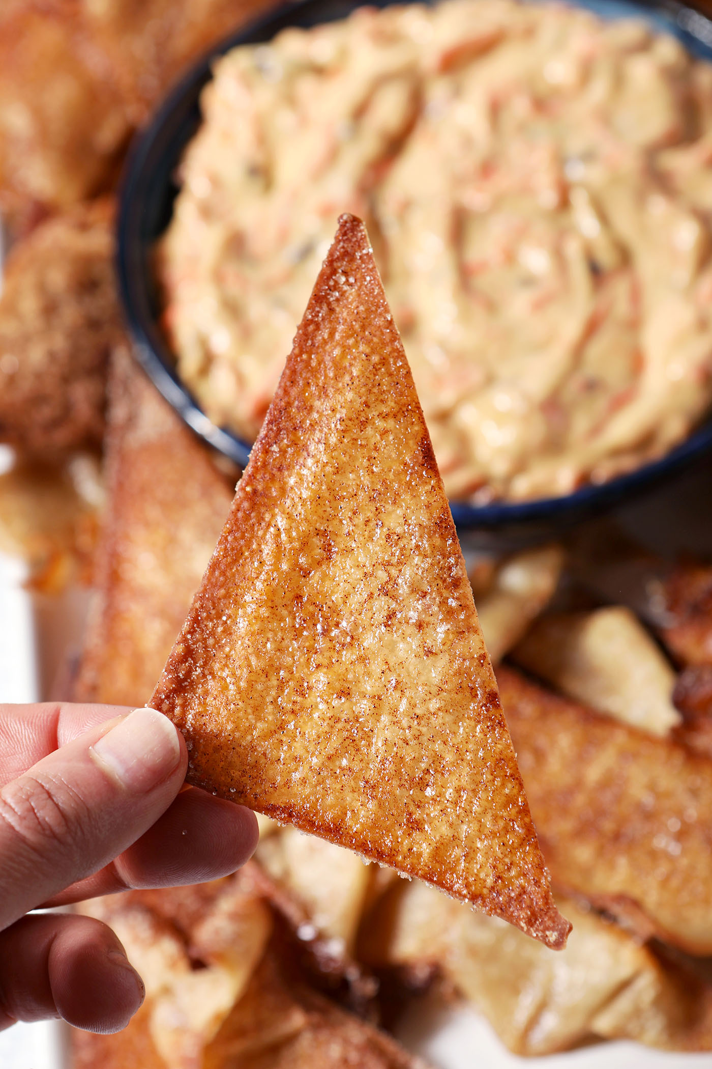 A hand holds a triangular Cinnamon Sugar Wonton Chip about a platter of more chips and carrot cake dip