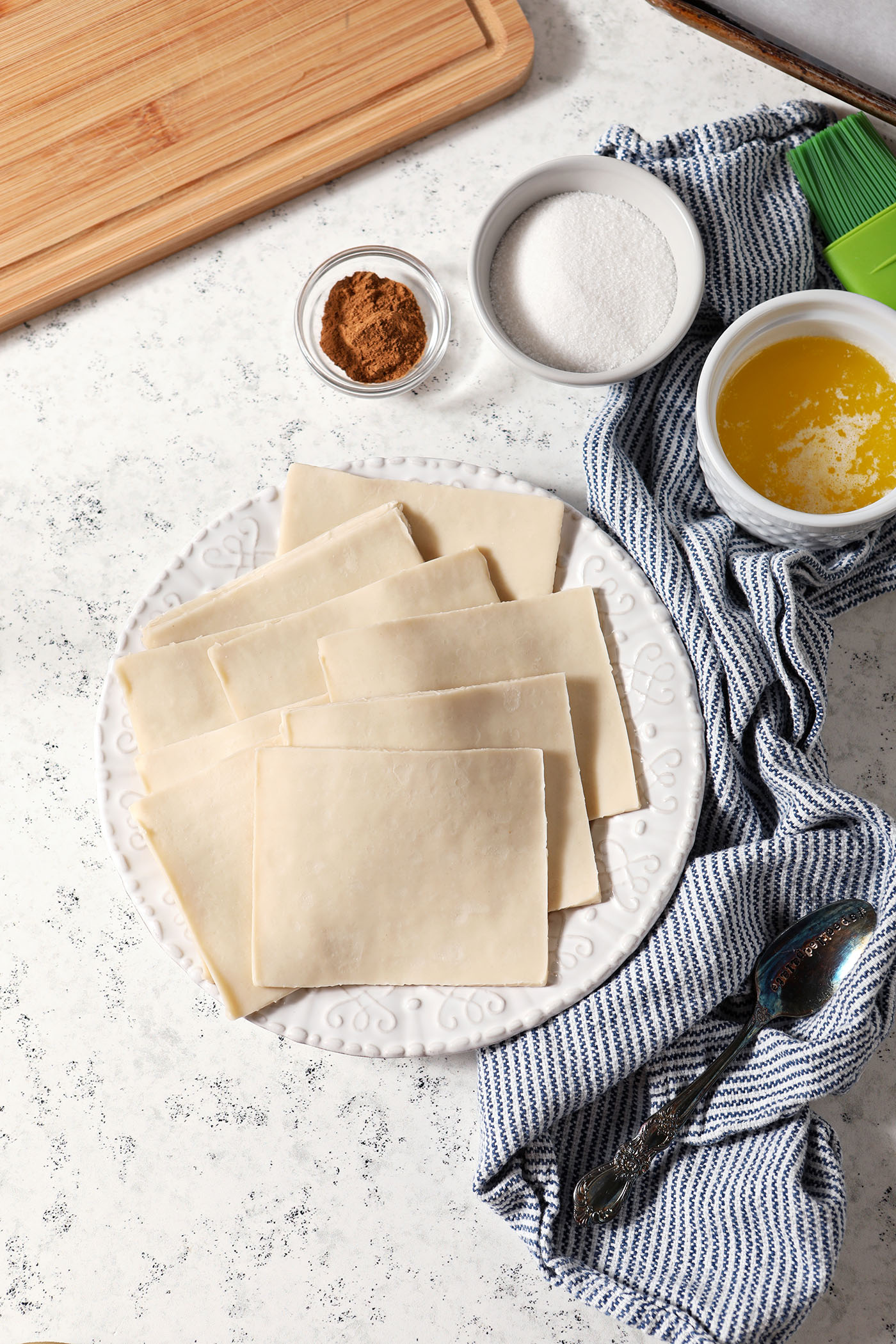 Wontons on a plate with bowls of cinnamon, sugar and butter surrounding them on a stone surface with a blue striped linen