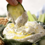 A cucumber dips into a bowl of Cottage Cheese Dip, surrounded by chips and veggies