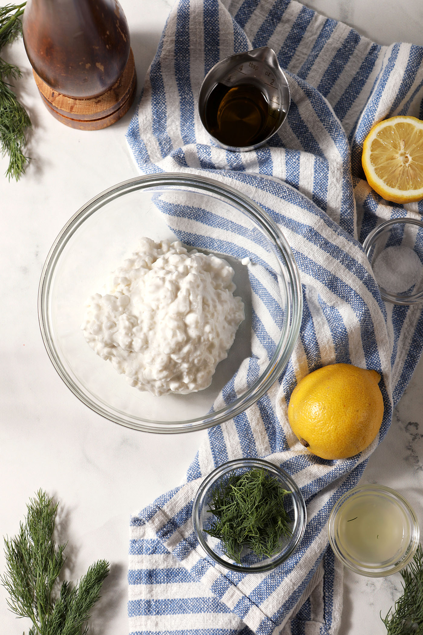 Cottage cheese in a bowl, surrounded by other ingredients in bowls, on a marble surface with a blue and white striped linen