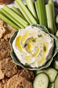 Overhead of Cottage Cheese Dip, garnished with dill, olive oil and lemon zest, on a platter with crackers, cucumber rounds and celery sticks