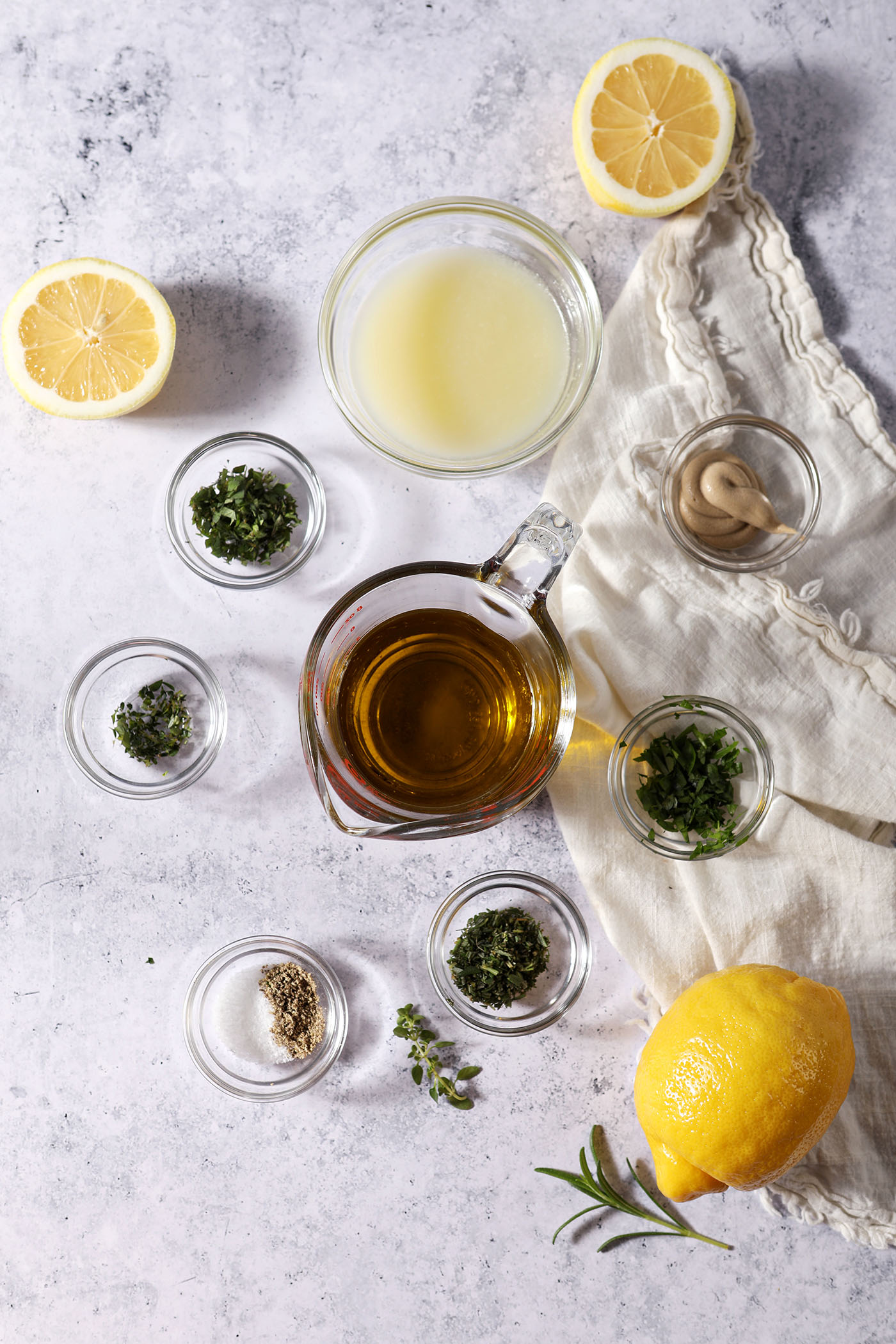 Ingredients to make a homemade lemon vinaigrette recipe in bowls and containers on a stone surface with a beige linen