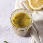 Homemade lemon vinaigrette in a Mason jar next to fresh herbs and a half lemon on a stone surface.