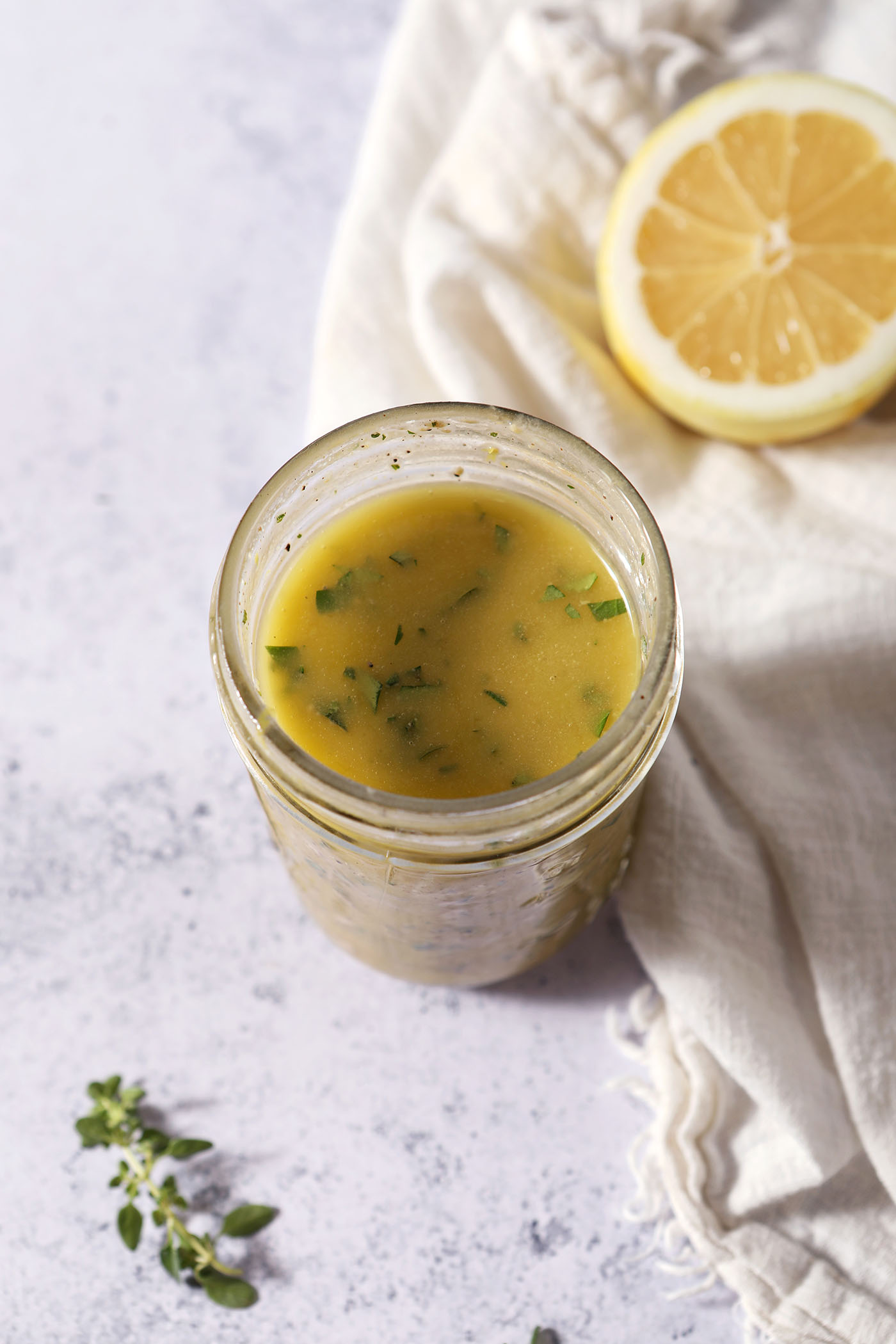 Homemade lemon vinaigrette in a Mason jar next to fresh herbs and a half lemon on a stone surface.