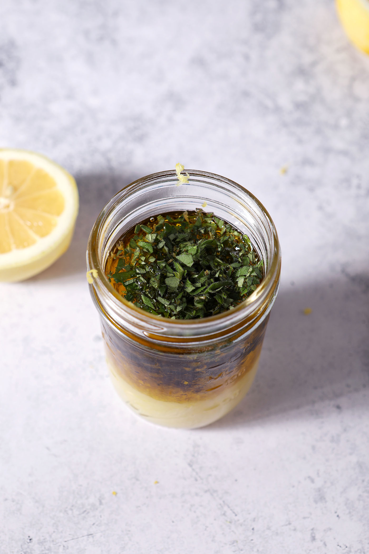 Fresh chopped herbs sit on top of other lemon vinaigrette ingredients in a mason jar on a stone surface next to a halved lemon before shaking