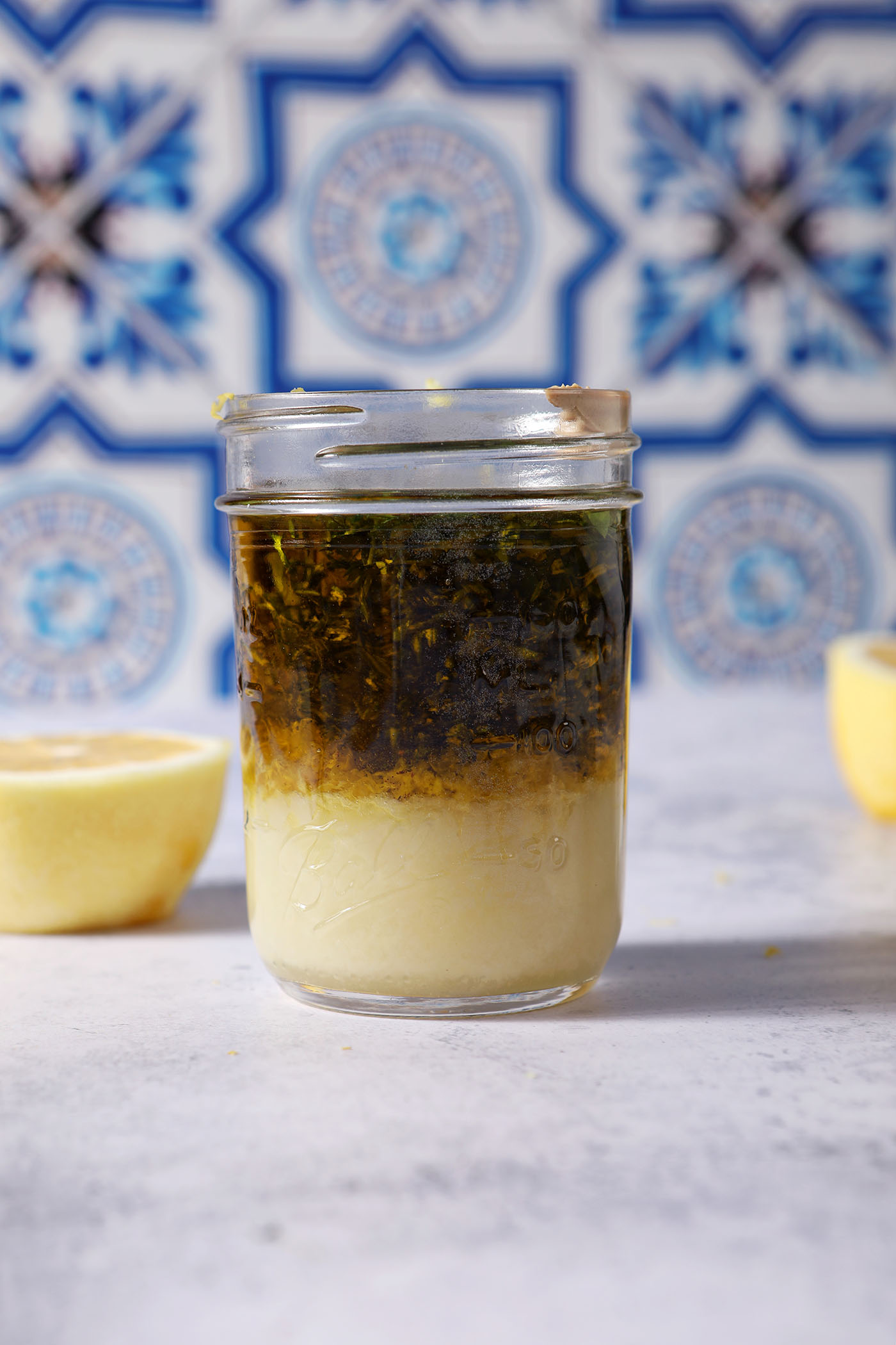A side view of fresh chopped herbs sit on top of other lemon vinaigrette ingredients in a mason jar on a stone surface next to a halved lemon before shaking