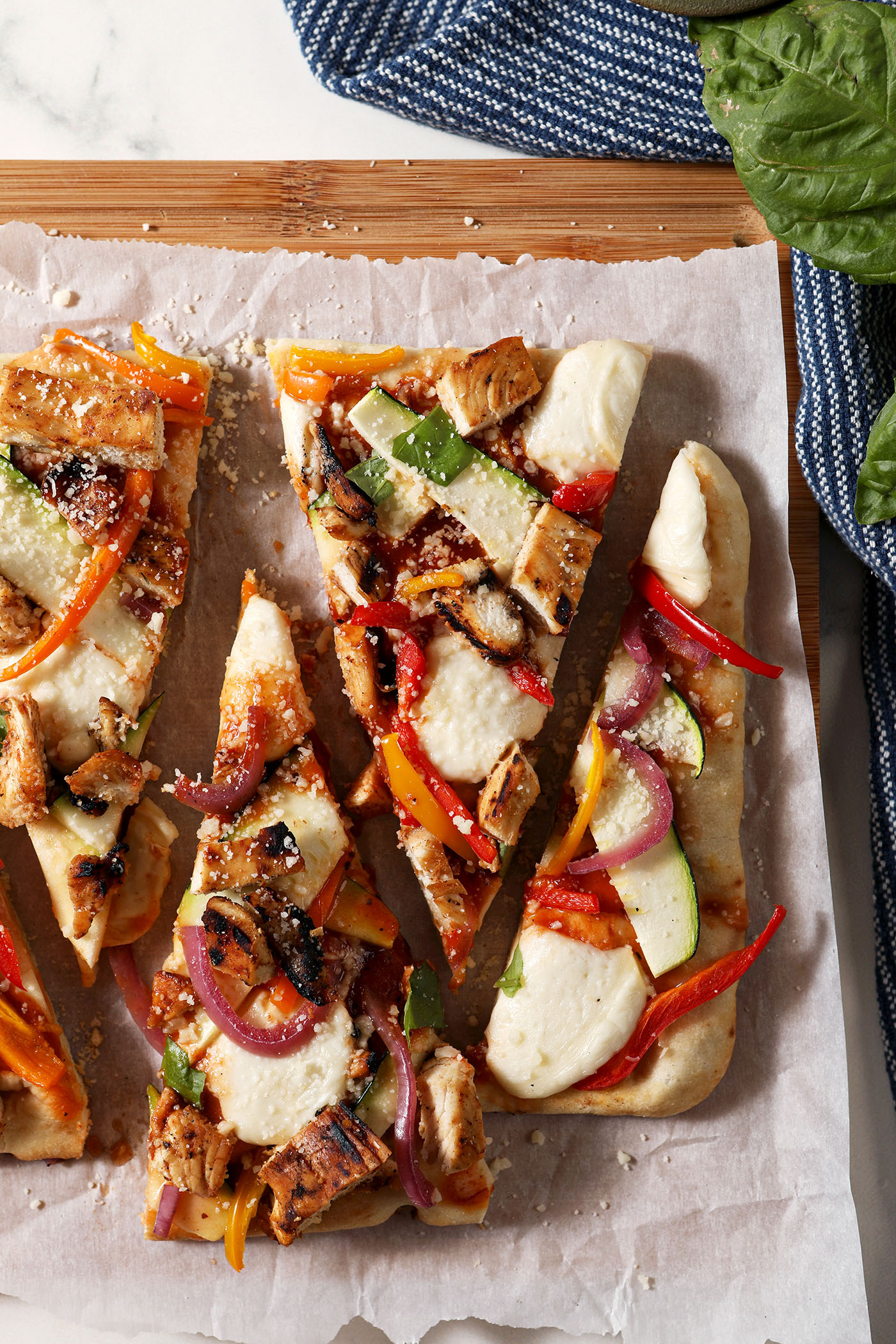 Sliced flatbread with barbecue chicken and vegetables on parchment paper on top of a wooden cutting board