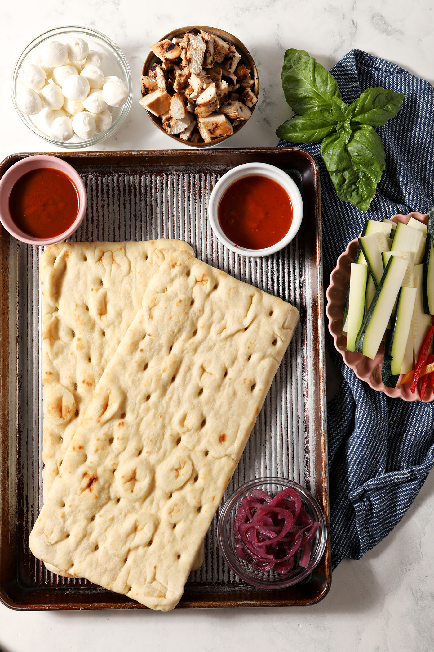 Two flatbreads on a sheet pan, surrounded by bowls of ingredients to make barbecue chicken flatbread, on a marble surface