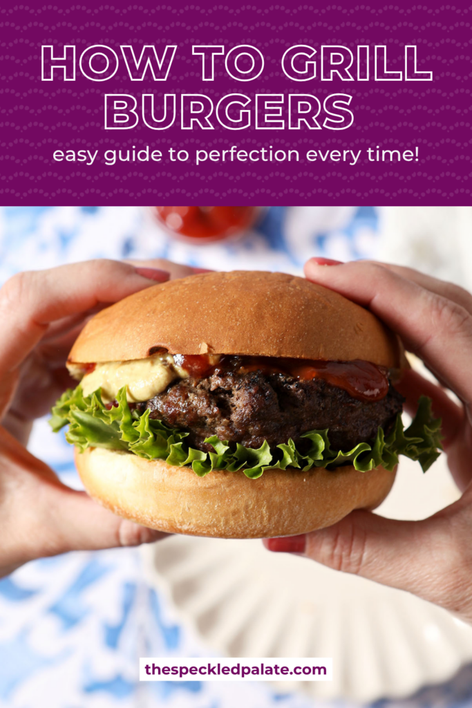 A woman holds a grilled burger in her hands above a blue and white tiled surface with plates, fries, and ketchup.