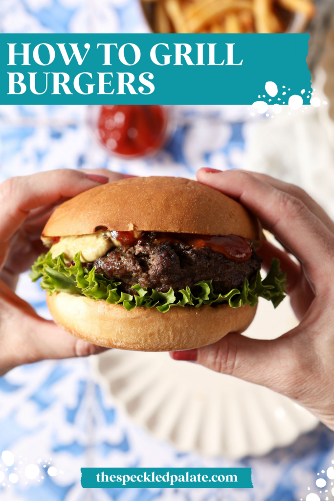 A woman holds a grilled burger in her hands above a blue and white tiled surface with plates, fries, and ketchup.