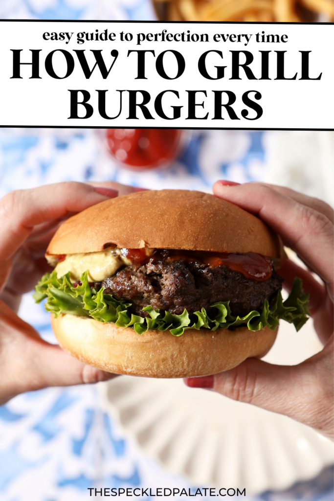 A woman holds a grilled burger in her hands above a blue and white tiled surface with plates, fries, and ketchup.