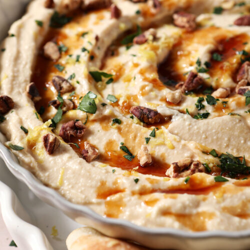 Close-up of hot honey dip with chopped herbs, hot honey, chopped pecans and lemon zest in a gray bowl.