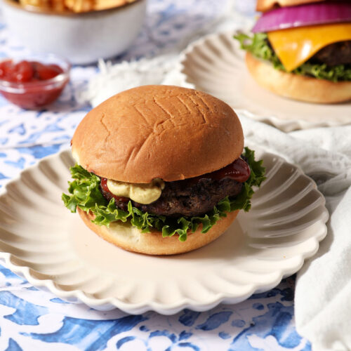 A grilled burger sits on a white plate, garnished with lettuce and seasonings, on a blue and white tiled surface next to other burgers, fries, and ketchup.
