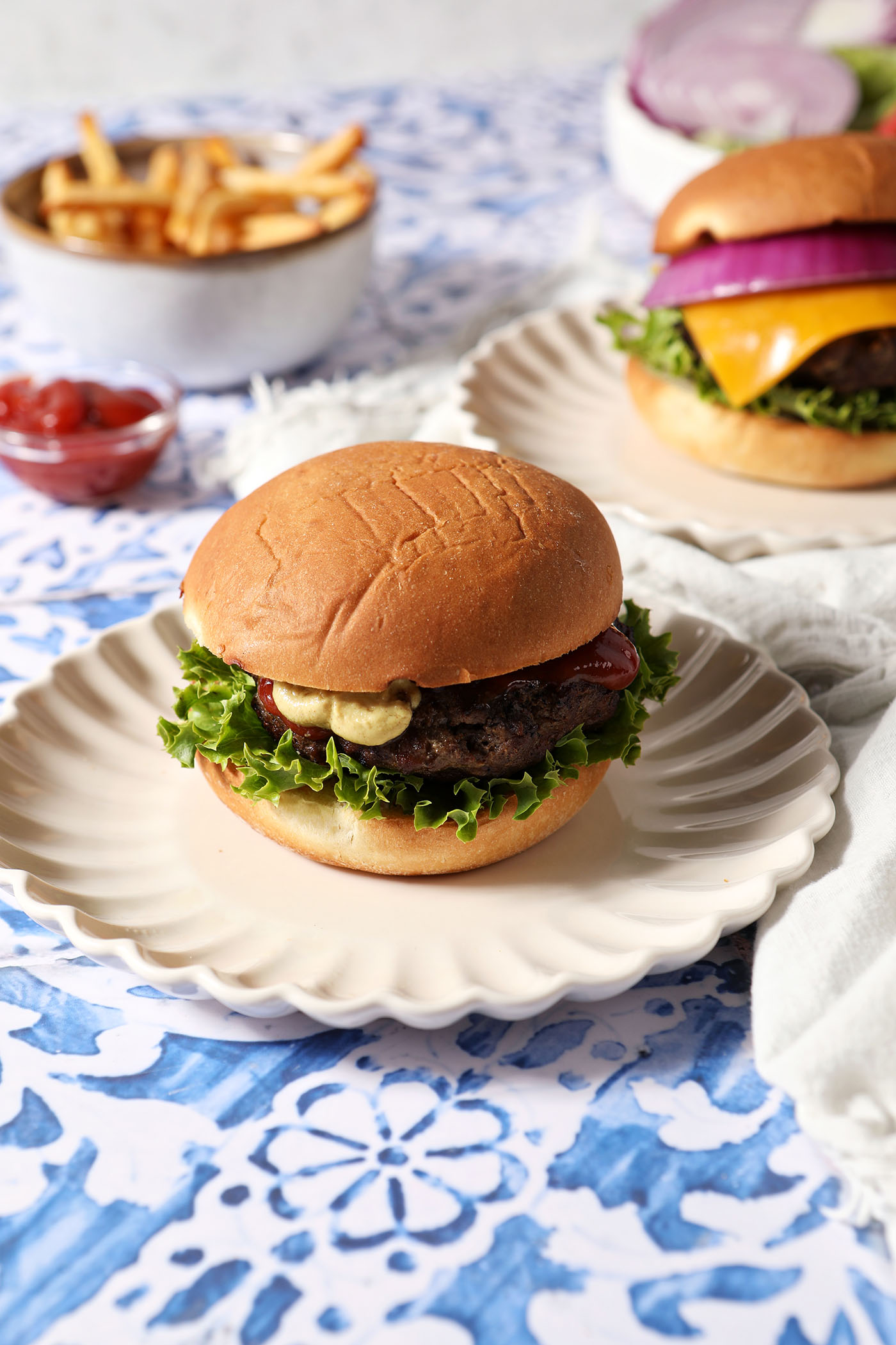 A grilled burger sits on a white plate, garnished with lettuce and seasonings, on a blue and white tiled surface next to other burgers, fries, and ketchup.