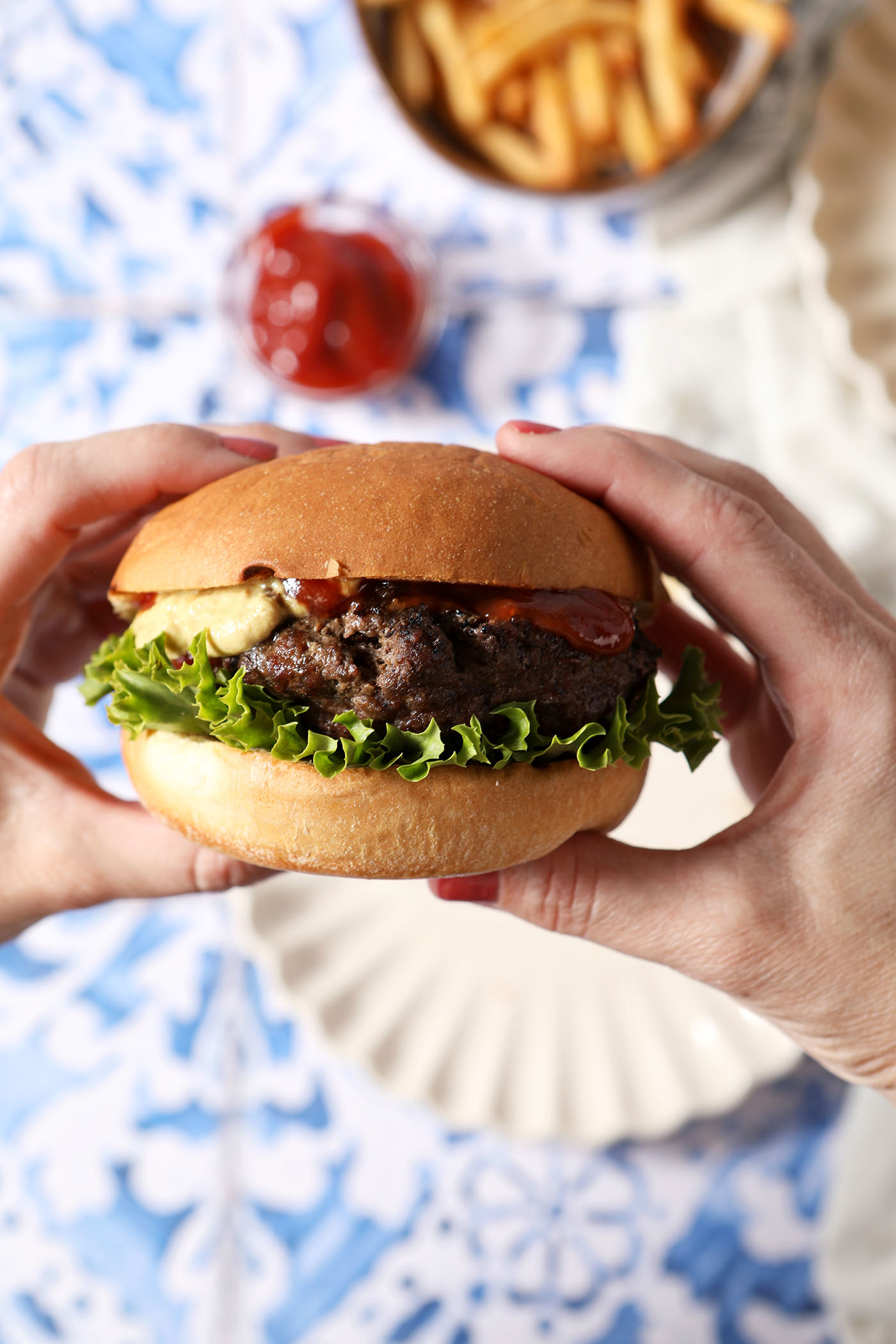 A woman holds a grilled burger in her hands above a blue and white tiled surface with plates, fries, and ketchup.