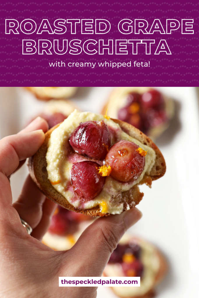 A hand holds a roasted grape crostini above a platter of more appetizers