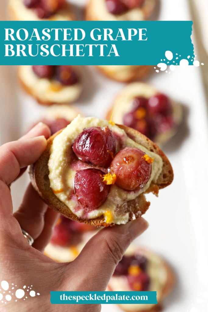 A hand holds a roasted grape crostini above a platter of more appetizers