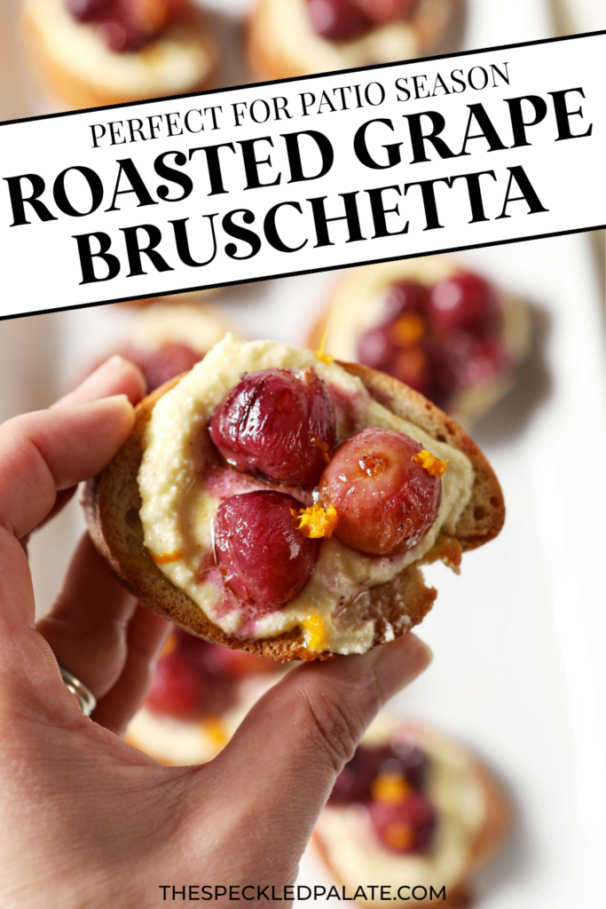 A hand holds a roasted grape crostini above a platter of more appetizers