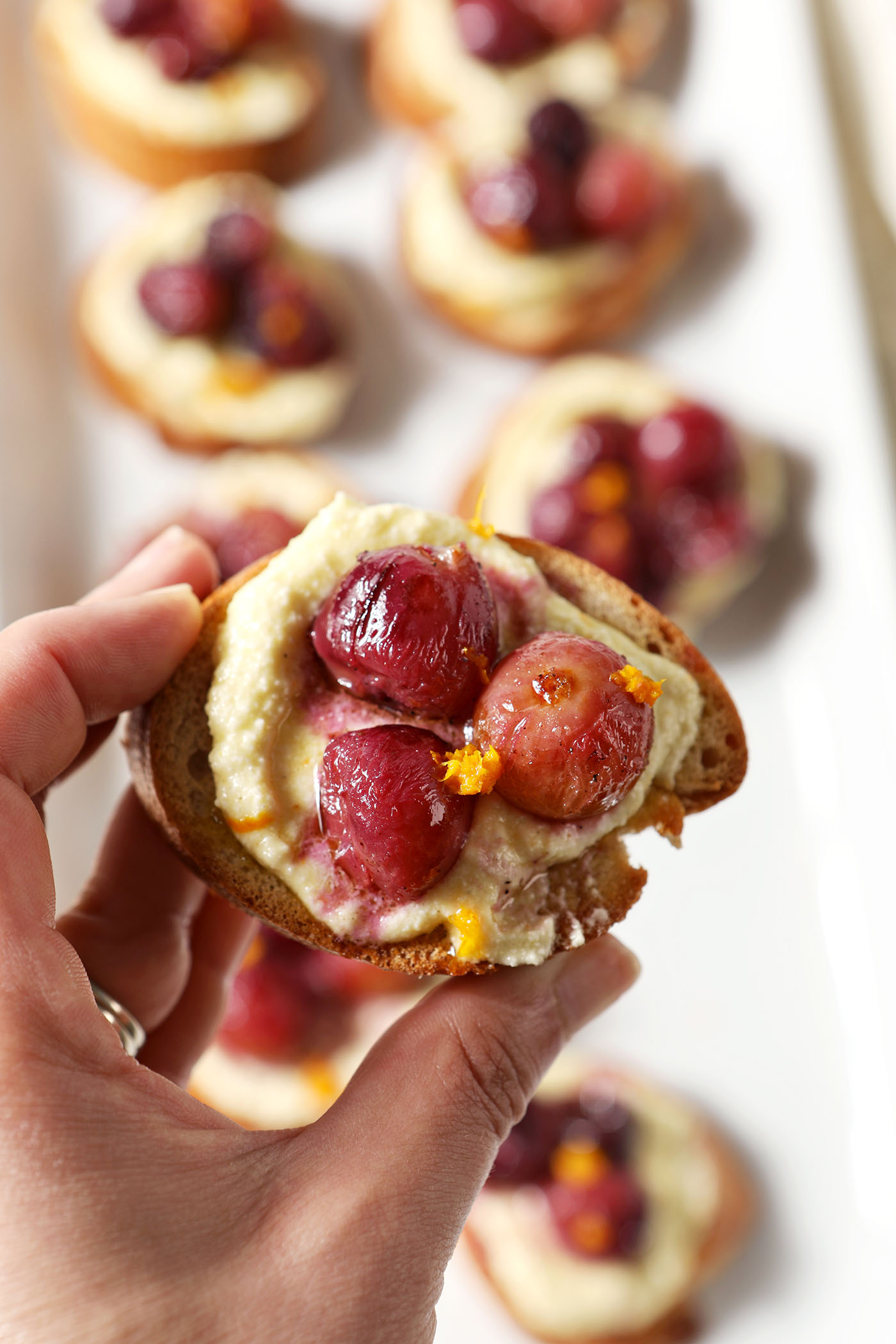 A hand holds a roasted grape crostini above a platter of more appetizers