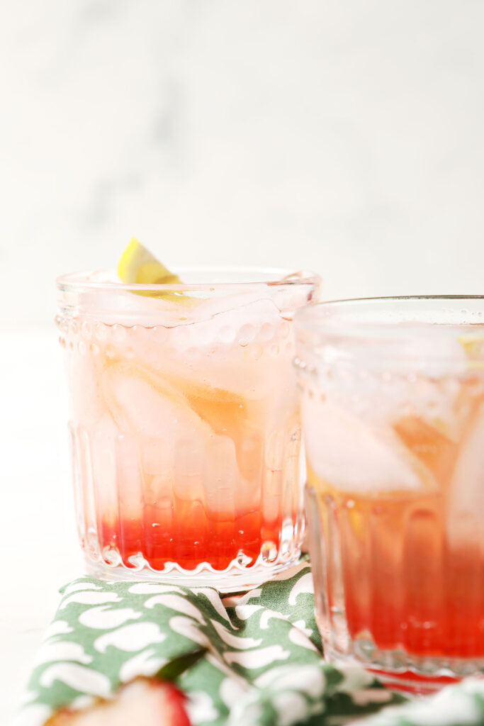 Side angle of two strawberry shrubs, garnished with lemon wedges, on a green patterned linen
