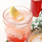 Close up of a strawberry shrub garnished with a lemon wedge next to another drink and a jar of shrub syrup
