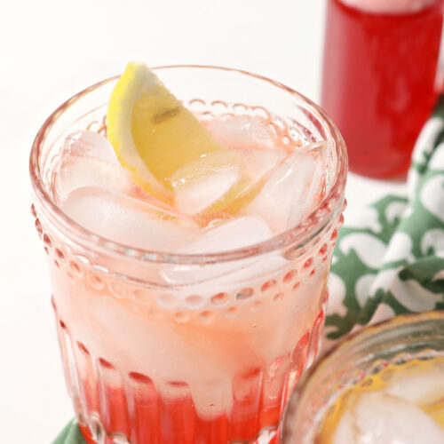 Close up of a strawberry shrub garnished with a lemon wedge next to another drink and a jar of shrub syrup
