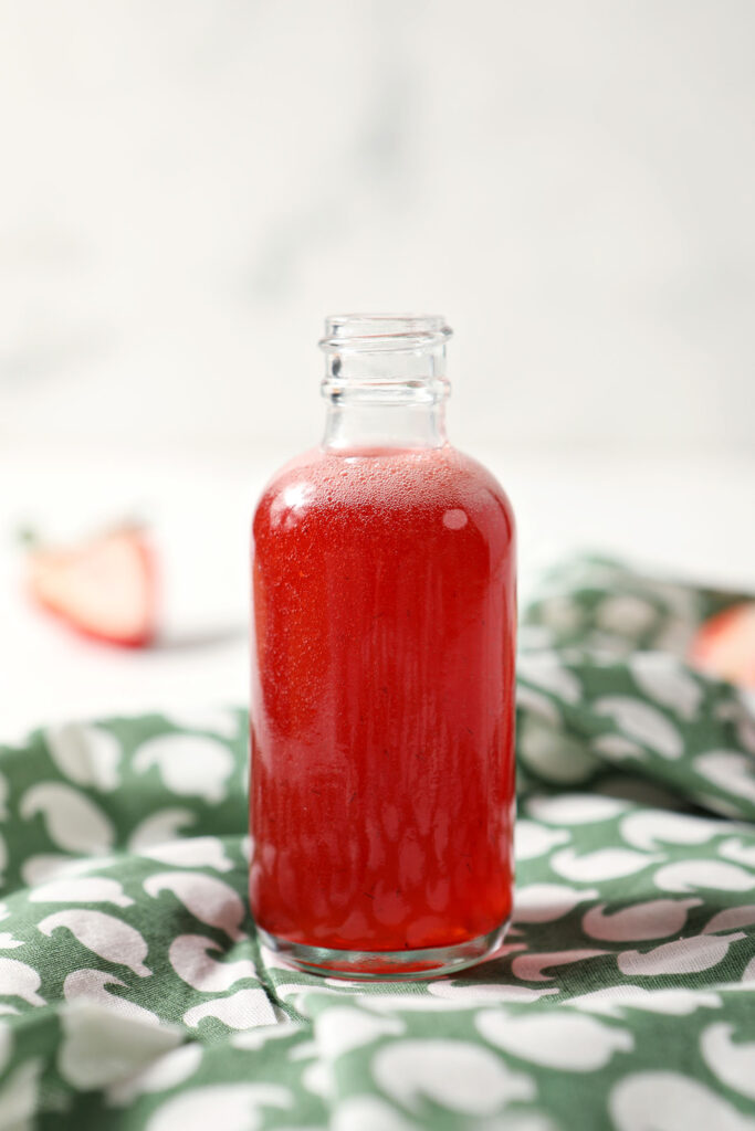A bottle of homemade strawberry shrub syrup on a green patterned linen