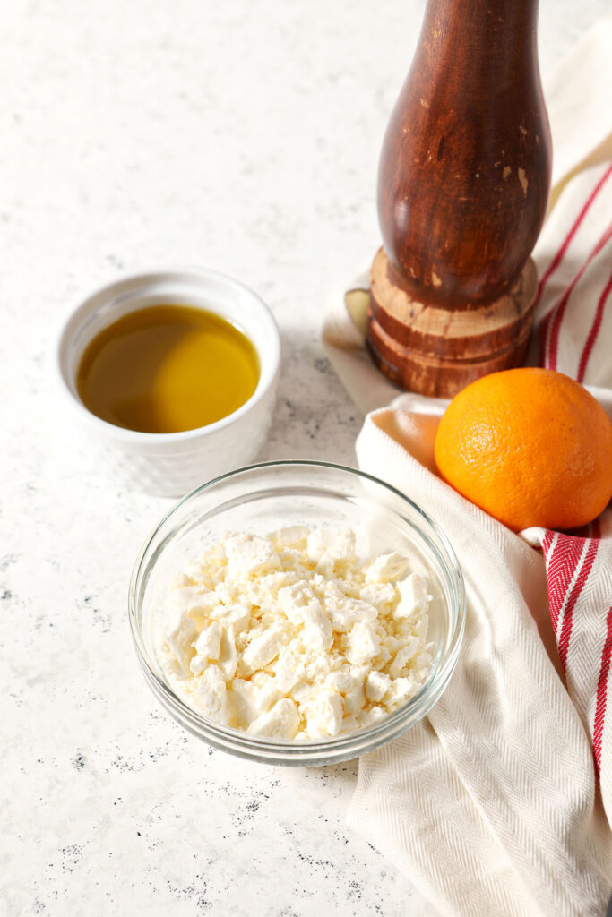 A bowl of feta with olive oil, an orange and black pepper on a white stone surface with a white and red striped linen