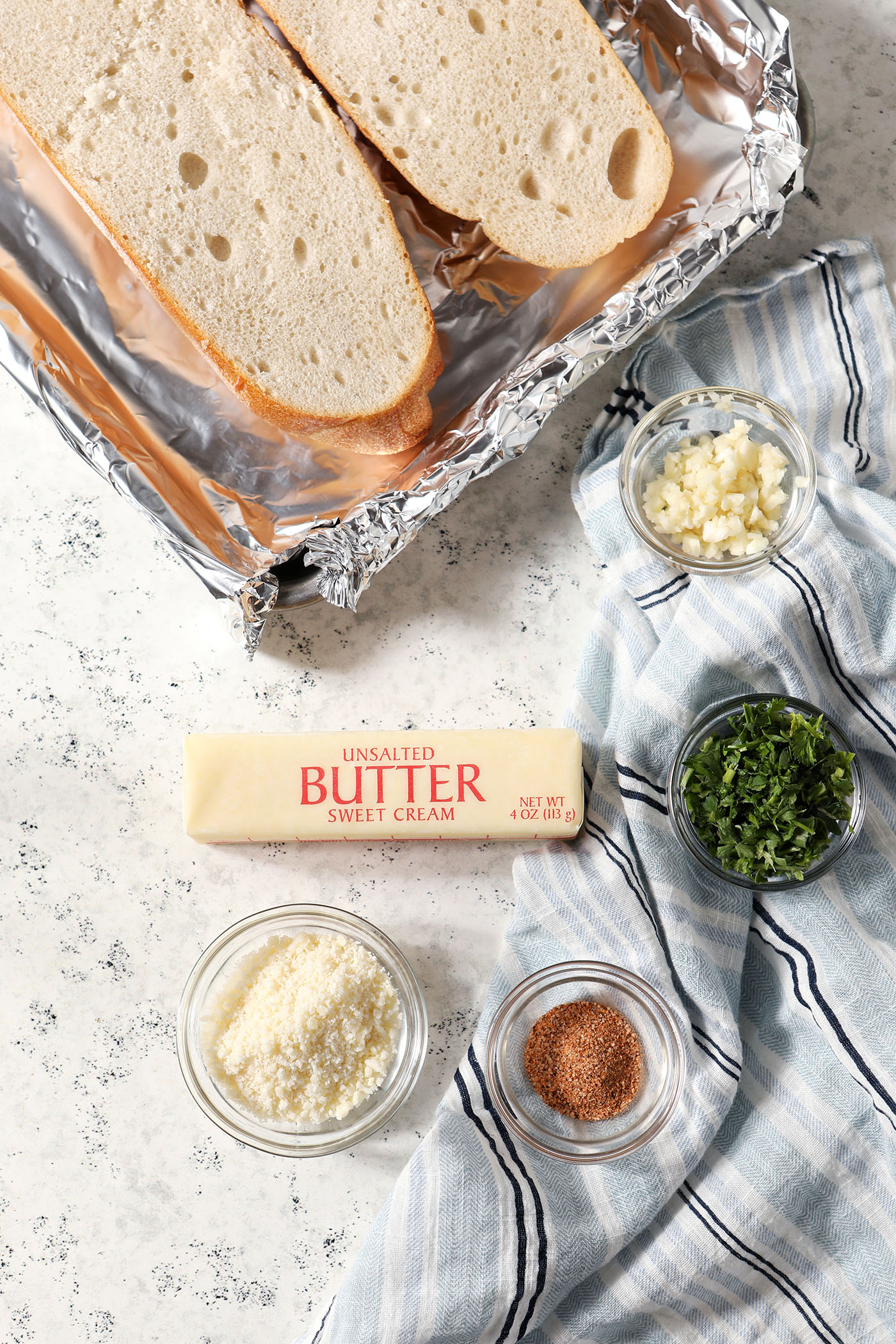 Ingredients in bowls to make Cajun garlic bread on a light stone surface with a blue striped linen