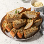 A plate holds several slices of Cajun garlic bread on a stone surface near bowls of Parmesan cheese and freshly minced parsley