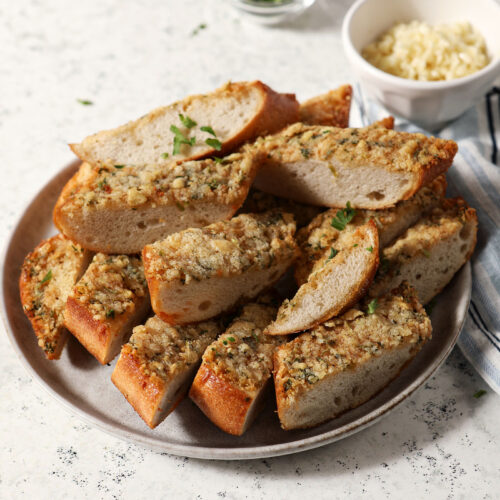 A plate holds several slices of Cajun garlic bread on a stone surface near bowls of Parmesan cheese and freshly minced parsley