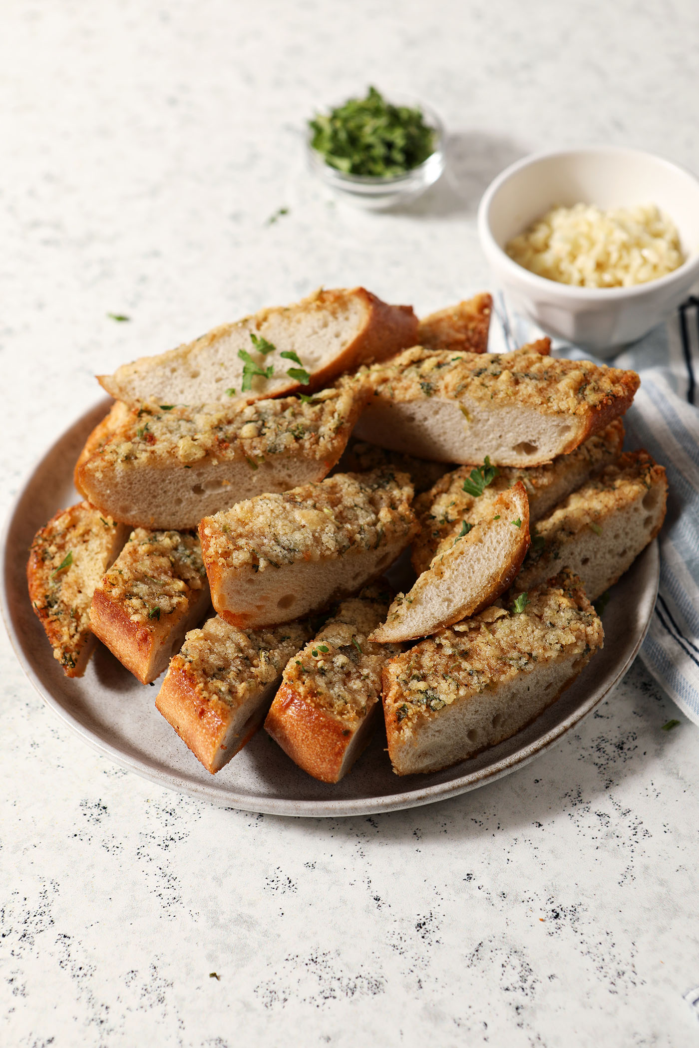 A plate holds several slices of Cajun garlic bread on a stone surface near bowls of Parmesan cheese and freshly minced parsley