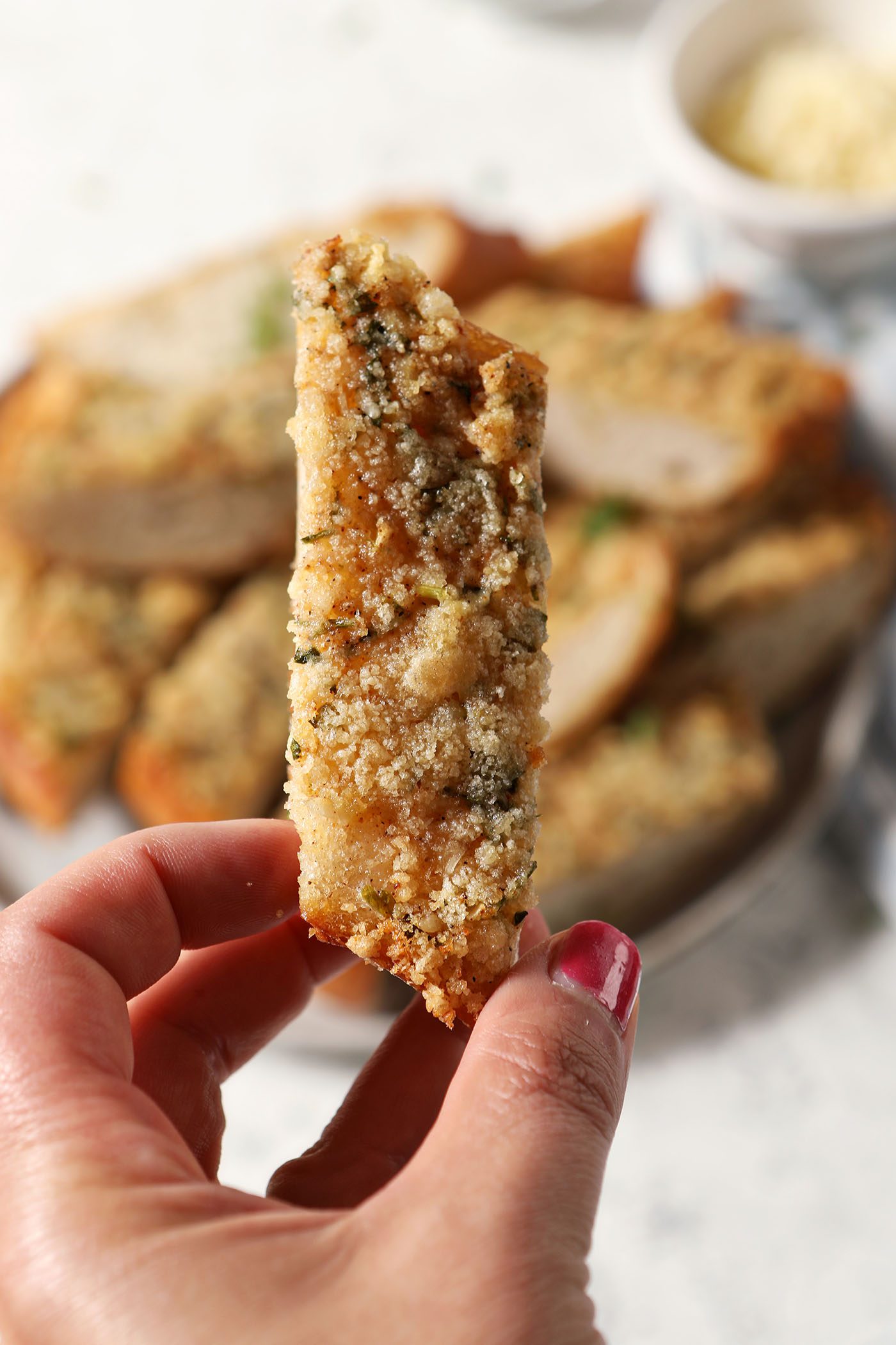 A hand with polished nails holds up a slice of homemade garlic bread in front of a platter of more bread